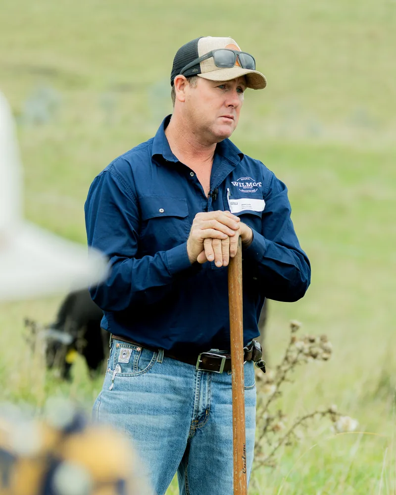 Shane Curtis, Group Operations Manager at Wilmot Cattle Co., leaning on a spade in a paddock with Angus cattle behind him